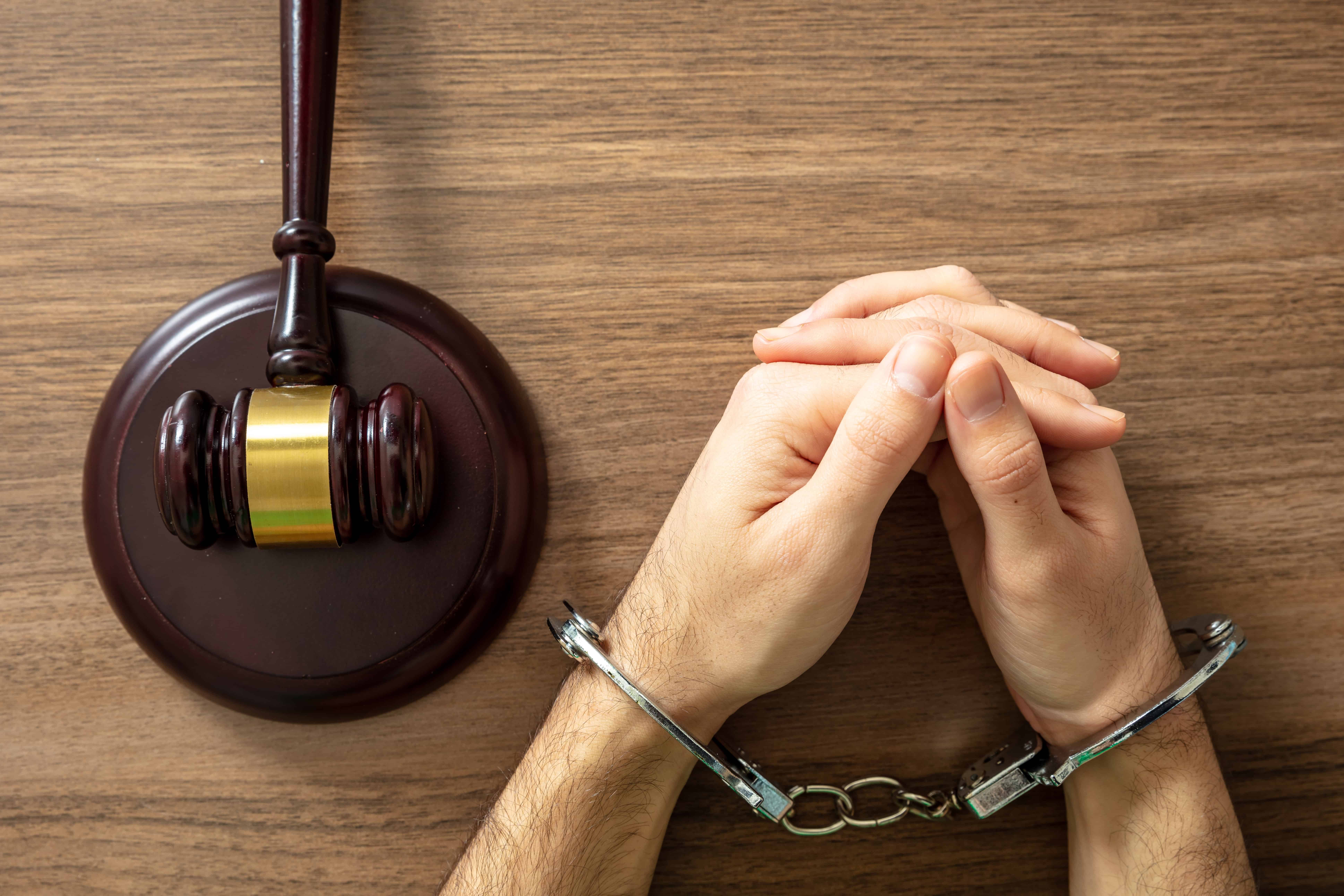 Handcuffed hands resting on a table beside a judge’s gavel, representing arrest, criminal charges, or the justice system.