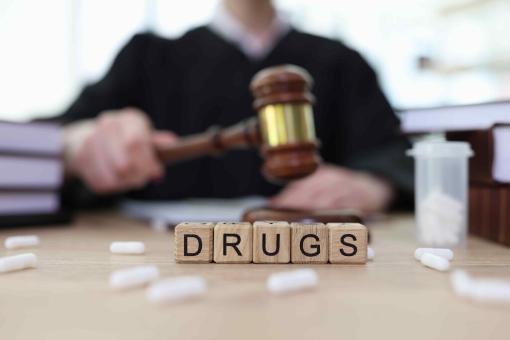 Judge holding a gavel behind the word ‘drugs’ spelled with blocks, representing drug-related criminal or legal cases.