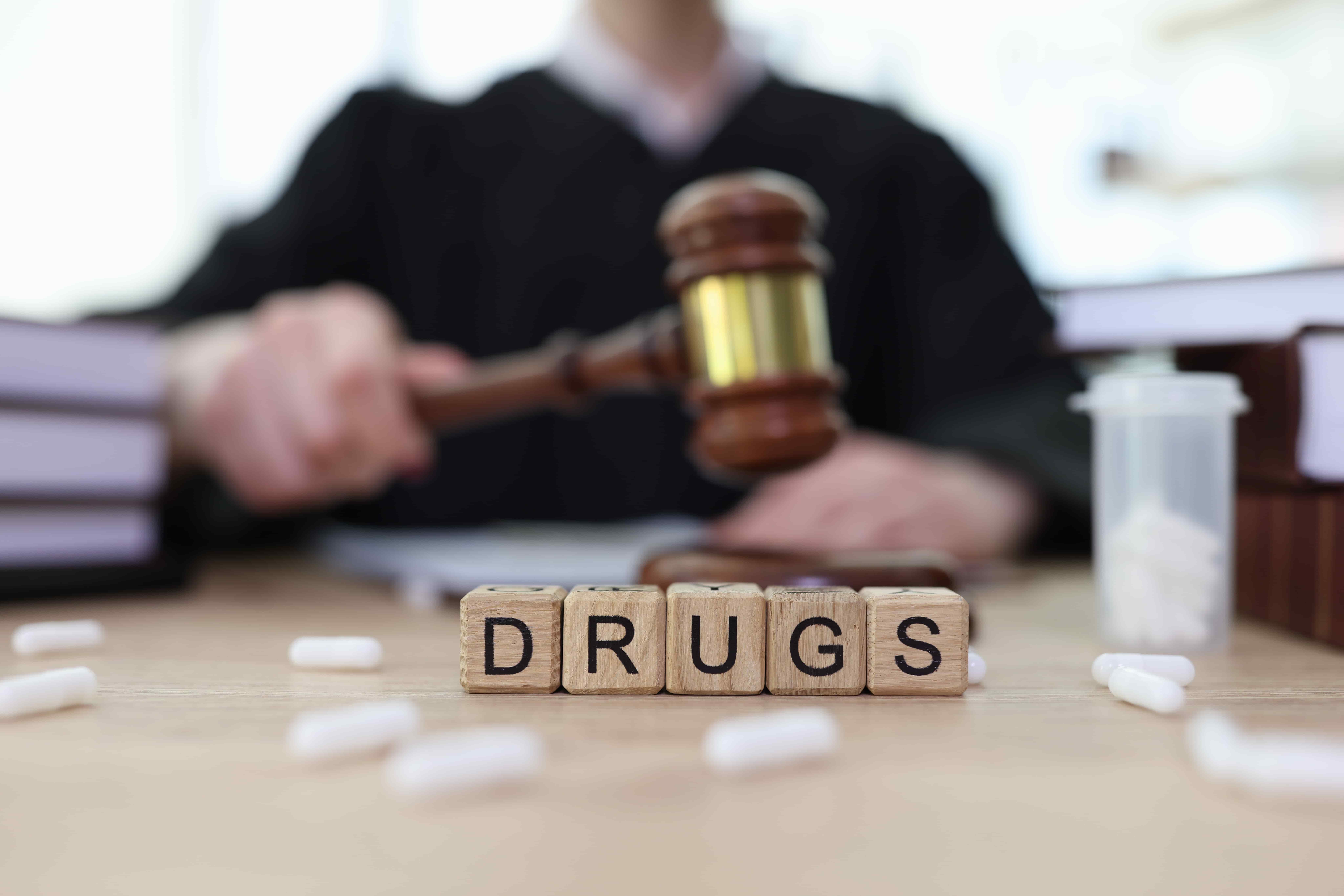 Judge holding a gavel behind the word ‘drugs’ spelled with blocks, representing drug-related criminal or legal cases.