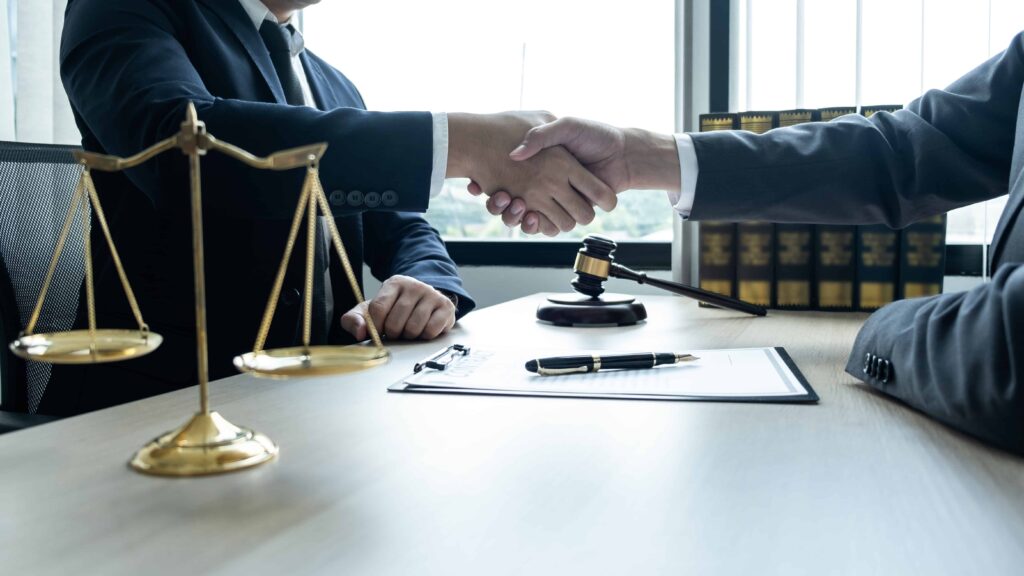 Lawyers shaking hands across a desk with a judge’s gavel and scales of justice, symbolizing a legal agreement or settlement.